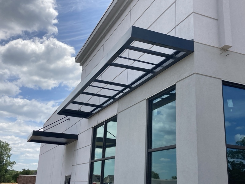a beautifully engineered custom cantilevered canopy and background overhead support canopy with hanger rods against a bright cloudy blue sky on a white building, crafted by the shade industry experts at Awnings by Bigley & Hogshire