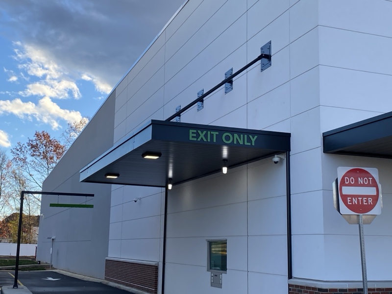A precision-crafted overhead support canopy on a white building with the blue sky and trees in the background, crafted by the shade industry experts, Awnings by Bigley and Hogshire.