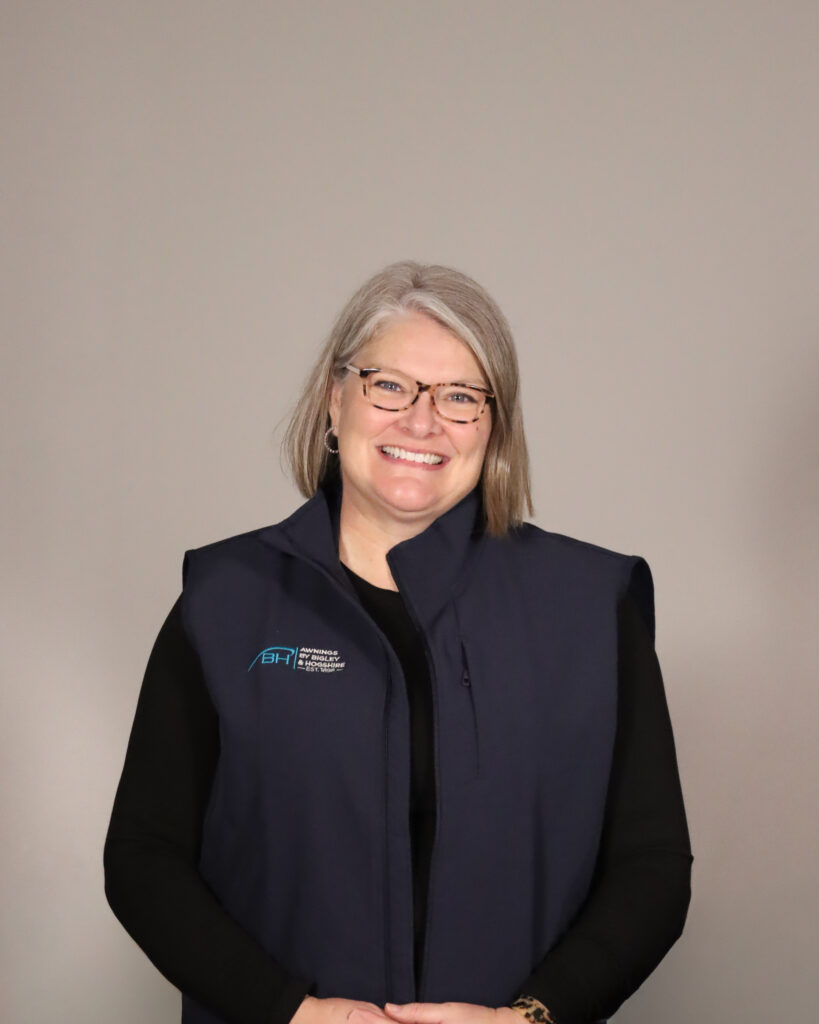 Joanna Bigley, Wife of the President and Human Resources Director for Awnings by Bigley & Hogshire stands with her hands folded, wearing a warm smile and a striking navy vest with the company logo on the front.